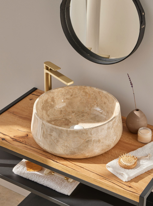 Ronde natuursteen waskom 40x40x15cm in warme beige tint op licht houten blad met gouden kraan, sfeervol gefotografeerd in moderne badkamer.