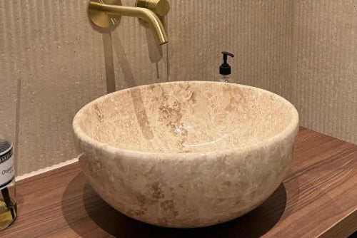 Zandkleurige natuurstenen waskom op houten badmeubel met gouden inbouwkraan in moderne toiletruimte