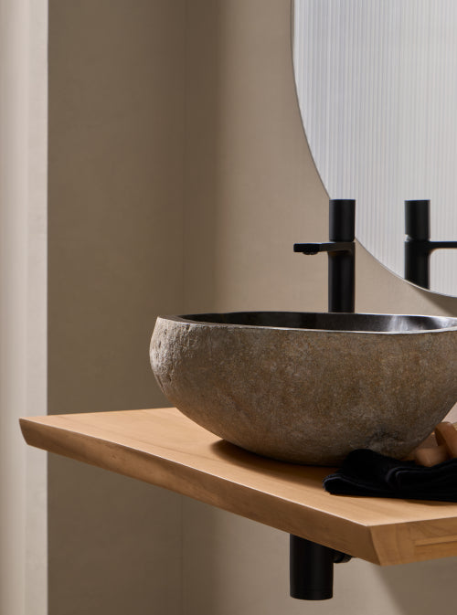 Natuurstenen riviersteen waskom in organische vorm op licht houten blad naast zwarte opbouwkraan 38x42cm